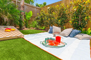 Sunny backyard picnic on green lawn with striped blanket, pitcher of red punch and cups on a tray, soda cans and cushions, a large wooden checkerboard game, tropical plants and a wooden fence