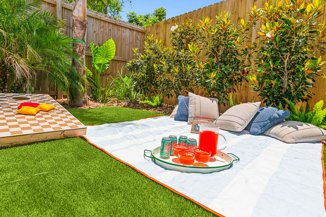 Sunny backyard picnic on green lawn with striped blanket, pitcher of red punch and cups on a tray, soda cans and cushions, a large wooden checkerboard game, tropical plants and a wooden fence