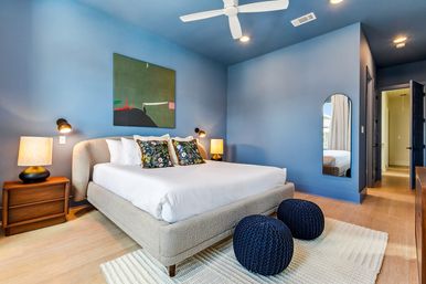 Modern blue master bedroom with king-size upholstered bed dressed in white linens and floral accent pillows, wooden nightstands with lamps, abstract wall art, white ceiling fan, two navy knit poufs on a cream rug, arched full-length mirror and light hardwood floors.