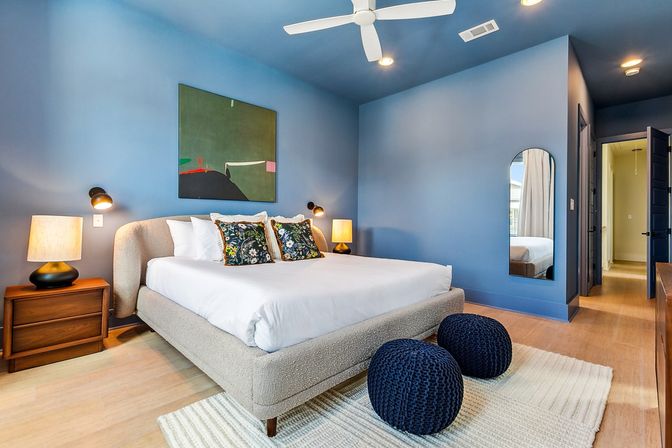 Modern blue master bedroom with king-size upholstered bed dressed in white linens and floral accent pillows, wooden nightstands with lamps, abstract wall art, white ceiling fan, two navy knit poufs on a cream rug, arched full-length mirror and light hardwood floors.
