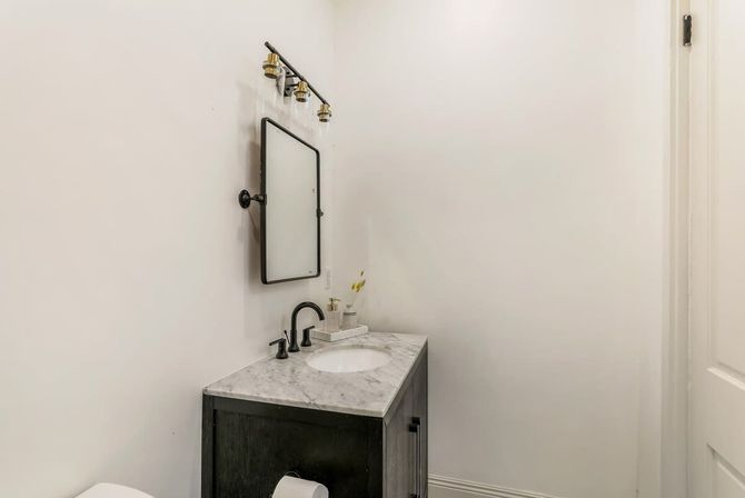 Chic, bright powder room with marble-top vanity, matte-black faucet and fixtures, rectangular wall mirror and brass-accented light fixture against crisp white walls.