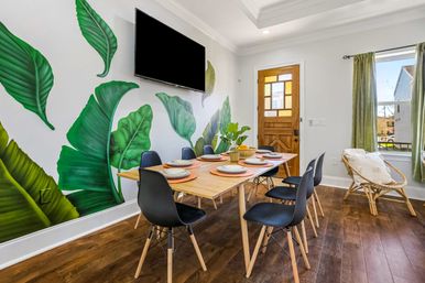 Bright dining room with wooden table set for six, black modern chairs and potted centerpiece, large vibrant tropical leaf mural on a white wall, wall-mounted TV, wooden front door, green-curtained window and hardwood floors.