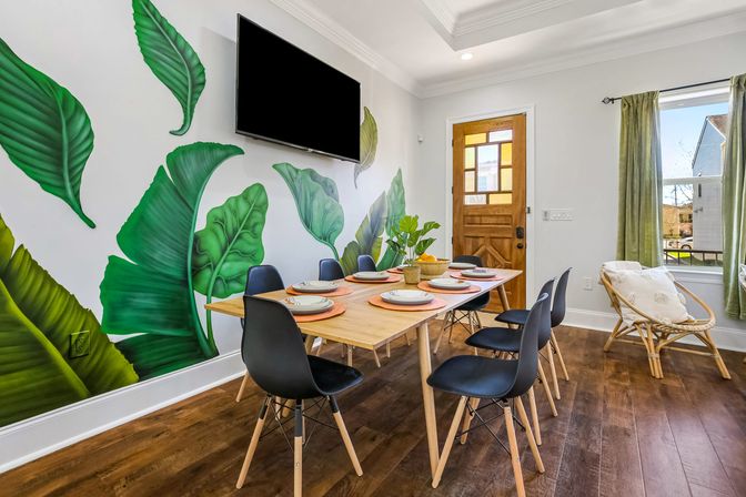 Bright dining room with wooden table set for six, black modern chairs and potted centerpiece, large vibrant tropical leaf mural on a white wall, wall-mounted TV, wooden front door, green-curtained window and hardwood floors.