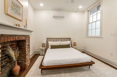 Bright minimalist bedroom with low wooden platform bed, white linens and green lumbar pillow, woven bedside lamps, exposed red-brick fireplace with vase of dried grasses, hardwood floors, neutral rug, sash window and wall-mounted AC — modern-meets-historic interior.