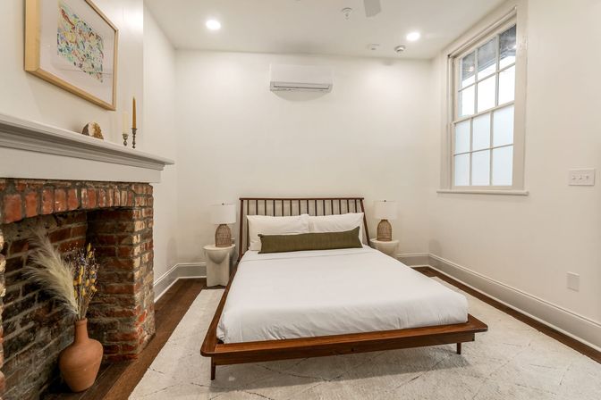 Bright minimalist bedroom with low wooden platform bed, white linens and green lumbar pillow, woven bedside lamps, exposed red-brick fireplace with vase of dried grasses, hardwood floors, neutral rug, sash window and wall-mounted AC — modern-meets-historic interior.