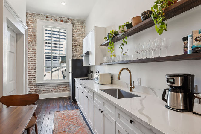 Sunlit urban loft galley kitchen with exposed brick wall, large window with city view, white cabinets and marble-look quartz countertops, brass faucet, open wood shelves with plants and glassware, dark hardwood floors and coffee maker.