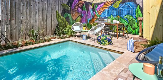 Sunny small backyard pool with turquoise water, tiled patio with round lounge chairs, dining table, BBQ and tropical plants in front of a vibrant mural on a wooden fence.
