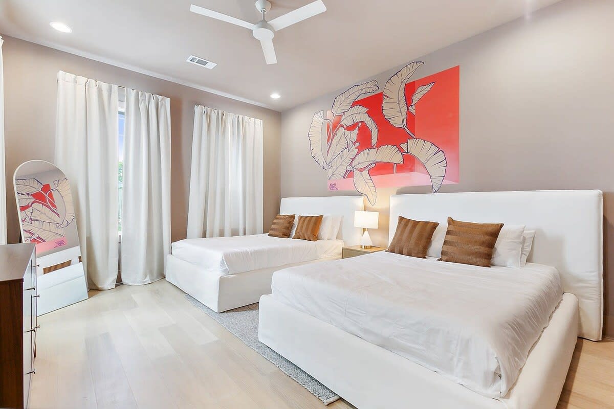 Bright modern bedroom interior with two white platform beds, tropical red leaf mural, white curtains, brown accent pillows, light wood floors and ceiling fan