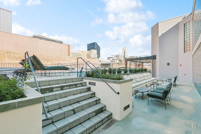 Sunny downtown rooftop terrace with cushioned sofa seating, lounge chairs, planter boxes and steps, overlooking a city skyline under a bright blue sky