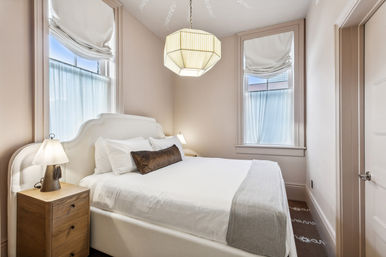 Light-filled bedroom interior with white upholstered headboard, crisp bedding, wooden nightstand and lamp, geometric pendant light, and two tall windows with sheer curtains and roman shades — cozy neutral-toned retreat.