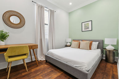 Bright, cozy bedroom with sage-green accent wall, gray upholstered bed with white linens and striped pillows, matching bedside lamps on round tables, wooden desk with a chartreuse mid-century chair, rattan wall mirror, potted plant, and warm hardwood floors.