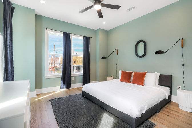 Bright modern urban bedroom with sage-green walls, low-profile gray upholstered bed with white linens and rust-orange pillows, black wall-mounted reading lamps and ceiling fan, large window with dark curtains showing a city construction view, hardwood floors and dark area rug.