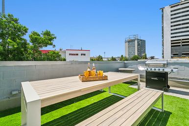Sunny urban rooftop terrace with wooden picnic table and benches on artificial turf, tray of drinks, a stainless-steel BBQ grill, low concrete wall and city skyline under a clear blue sky.