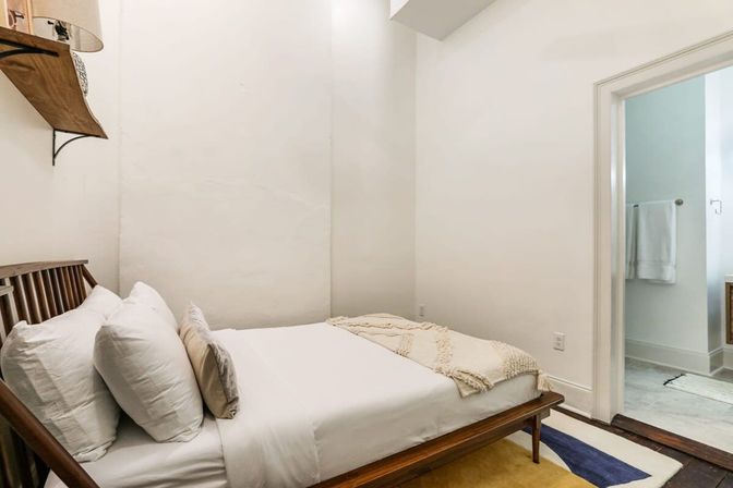 Cozy minimalist bedroom with a mid-century wooden bed, white linens and pillows, textured cream throw and area rug, neutral walls and an open doorway to a bright bathroom.