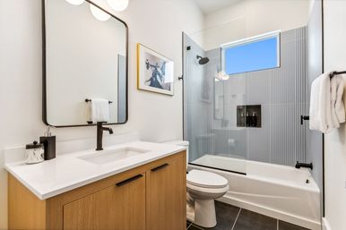 Bright modern bathroom with wood vanity and white countertop, matte-black faucet, frameless glass shower/tub with vertical gray tile and high window.
