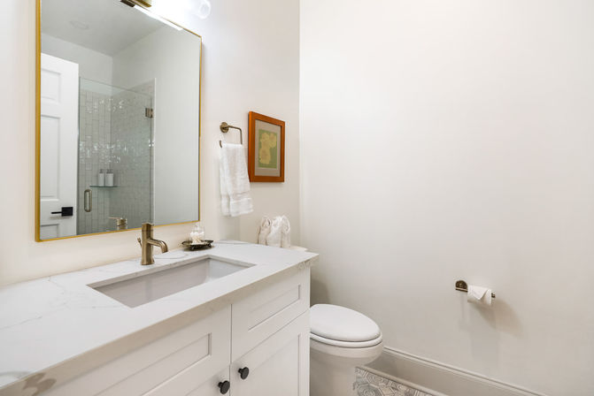 Bright modern residential powder room with white vanity and marble countertop, brass faucet, gold-framed mirror reflecting a glass shower, wall towel and framed art, patterned tile floor and toilet.
