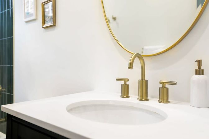 Sleek modern bathroom vanity with a white countertop and oval sink, brushed brass faucet and matching handles, round gold-framed mirror and white soap dispenser.