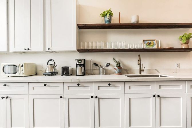 Bright modern white kitchen with marble countertop and open wood shelves, countertop appliances (microwave, kettle, toaster, coffee maker), potted plants, and stainless steel sink