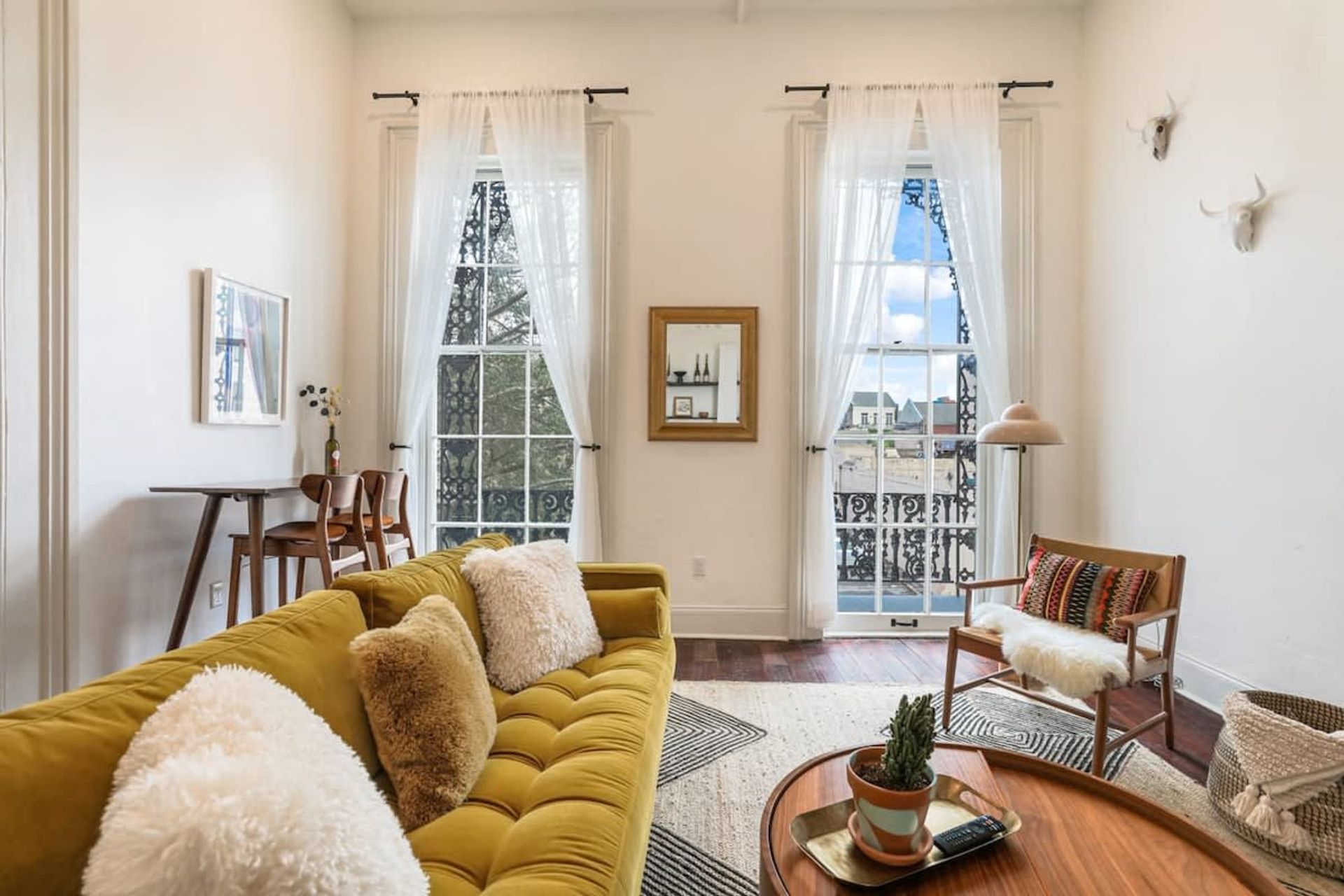 Sunlit modern living room with a mustard-yellow tufted sofa, sheer-curtained tall windows opening to a wrought-iron balcony, mid-century wooden chair with colorful pillow, round wood coffee table with potted plant, and hardwood floors.