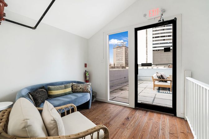Cozy urban loft lounge with a blue curved sofa and rattan armchair on wood floors, glass door opening to a sunlit rooftop terrace with downtown high-rise views.