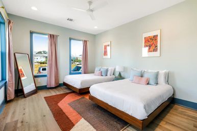 Airy modern guest bedroom with two wood platform beds in white linens and pastel throw pillows, colorful geometric rug, full‑length leaning mirror, three tall windows with blush curtains, light wood floors, ceiling fan and framed wall art.