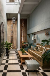 Sunlit narrow high-ceiling living room with black-and-white checkered tile, velvet brown sofa with patterned cushions, teal ottoman, wooden coffee table, tall gold drapes, hanging lantern and potted plants.