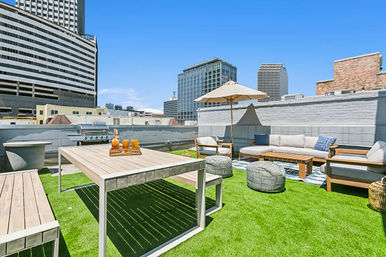 Sunny urban rooftop patio with artificial grass, modern dining table, lounge sofa, umbrella, poufs and downtown skyscrapers in the background.