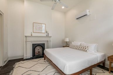 Bright, cozy minimalist bedroom with white walls, wooden platform bed dressed in crisp white linens and patterned lumbar pillow, vintage fireplace mantel, cream geometric rug, bedside tables with lamps, ceiling fan and wall-mounted AC.