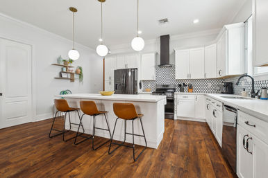 Bright modern white kitchen with large island, three brown leather bar stools, globe pendant lights, dark hardwood floors, stainless appliances and black-and-white geometric tile backsplash.