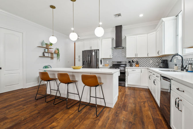 Bright modern white kitchen with large island, three brown leather bar stools, globe pendant lights, dark hardwood floors, stainless appliances and black-and-white geometric tile backsplash.