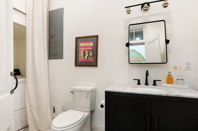 Bright modern bathroom with white walls, black vanity and marble countertop, black faucet and square mirror, white toilet, colorful framed New Orleans poster, overhead light fixture and stacked washer hidden behind a beige curtain.