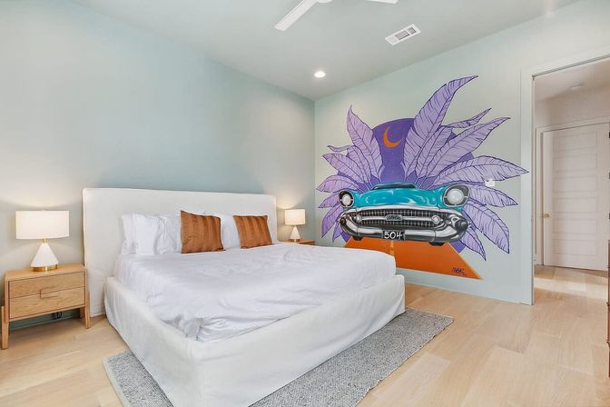 Bright modern bedroom with king-size white bed, light wood nightstands and lamps, pale hardwood floors, and a colorful wall mural of a turquoise vintage car framed by oversized purple tropical leaves and a crescent moon.