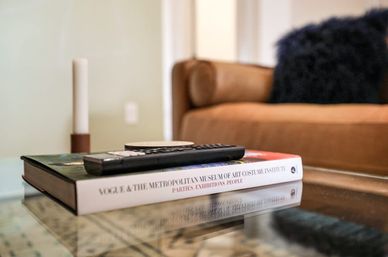 TV remote perched on a hardcover art book on a glass coffee table in a cozy modern living room with a tan sofa, fuzzy pillow, and candle in the background.