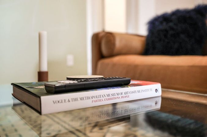 TV remote perched on a hardcover art book on a glass coffee table in a cozy modern living room with a tan sofa, fuzzy pillow, and candle in the background.
