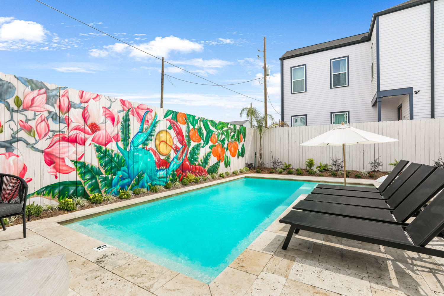 Urban residential backyard pool with turquoise water, black lounge chairs and a white umbrella on a stone patio, and a vibrant tropical mural of large flowers, fruits and a colorful bird painted on a wooden fence beside a modern two-story home.