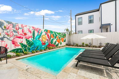 Urban residential backyard pool with turquoise water, black lounge chairs and a white umbrella on a stone patio, and a vibrant tropical mural of large flowers, fruits and a colorful bird painted on a wooden fence beside a modern two-story home.