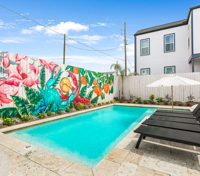 Urban residential backyard pool with turquoise water, black lounge chairs and a white umbrella on a stone patio, and a vibrant tropical mural of large flowers, fruits and a colorful bird painted on a wooden fence beside a modern two-story home.