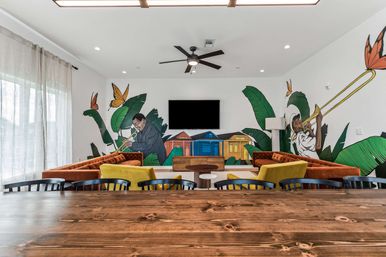 Bright modern community lounge with colorful jazz mural of trumpet and trombone players, velvet orange sofas, yellow accent chairs, wall-mounted TV, wooden table and tropical leaf motifs