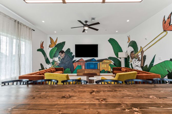 Bright modern community lounge with colorful jazz mural of trumpet and trombone players, velvet orange sofas, yellow accent chairs, wall-mounted TV, wooden table and tropical leaf motifs
