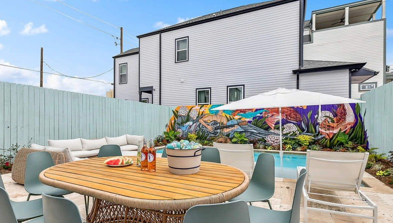 Sunlit backyard pool patio with round wooden dining table topped with watermelon and bottled drinks, teal chairs, white umbrella and lounge seating, vibrant ocean-themed mural on a pale blue fence in front of a modern gray townhouse.