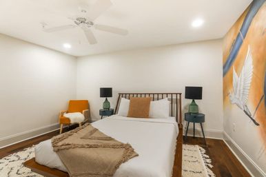 Cozy contemporary bedroom with wooden platform bed, crisp white linens and tan throw, mustard accent chair, green bedside lamps on round tables, hardwood floors, ceiling fan and a vibrant mural of a flying white bird.