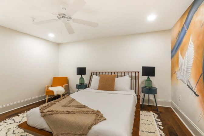 Cozy contemporary bedroom with wooden platform bed, crisp white linens and tan throw, mustard accent chair, green bedside lamps on round tables, hardwood floors, ceiling fan and a vibrant mural of a flying white bird.