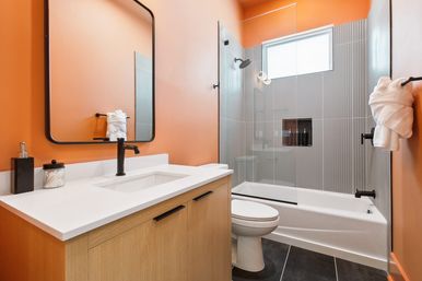 Bright modern bathroom interior with bold orange walls, white quartz vanity on light wood cabinet, black faucet and rectangular mirror, glass-enclosed bathtub-shower with vertical gray tile and high window, toilet and neatly folded towel on black hardware.
