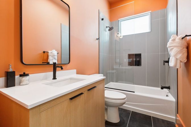 Bright modern bathroom interior with bold orange walls, white quartz vanity on light wood cabinet, black faucet and rectangular mirror, glass-enclosed bathtub-shower with vertical gray tile and high window, toilet and neatly folded towel on black hardware.