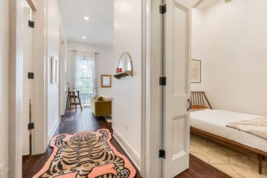 Light-filled apartment hallway with dark hardwood floors and a playful tiger-shaped rug, open doorway to a minimalist bedroom with wooden platform bed, and a living area with a mustard-yellow sofa and tall window.