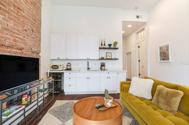 Bright open-plan studio living room with exposed brick wall, white kitchenette and floating shelves, mustard-green velvet sofa with fluffy pillows, round wooden coffee table with potted cactus, large flat-screen TV on metal console and patterned rug on dark hardwood floors.