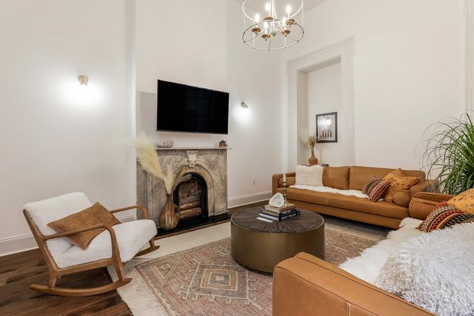 Bright boho-modern living room in an urban apartment with tan leather sofas, a cozy sheepskin rocking chair, marble fireplace with wall-mounted TV, round brass coffee table on a patterned rug, gold chandelier and potted plant.