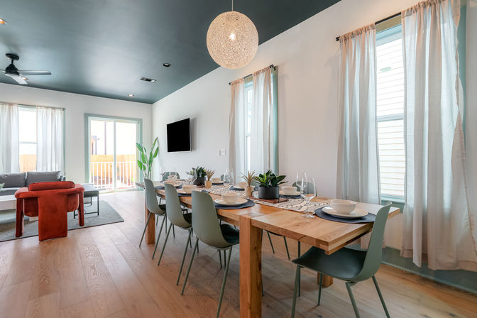 Bright modern open-plan dining and living area with a wooden dining table set for six, sage-green chairs, pendant light, hardwood floors, light curtains, and a sliding glass door to a sunny backyard.
