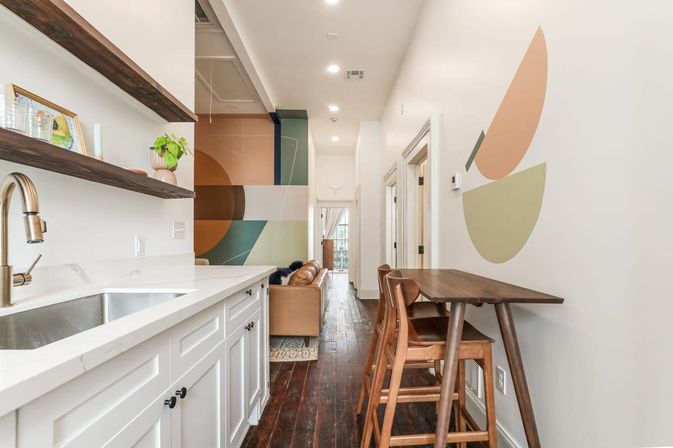 Bright modern narrow apartment kitchen and hallway with white shaker cabinets, brass faucet, wood floating shelves, geometric wall art, wooden bar table and stools, leather sofa and dark hardwood floors.