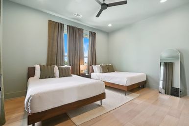 Bright modern guest bedroom with two wooden platform beds, white linens and green-brown pillows, brown curtains, bedside lamp, ceiling fan, and arched floor mirror.